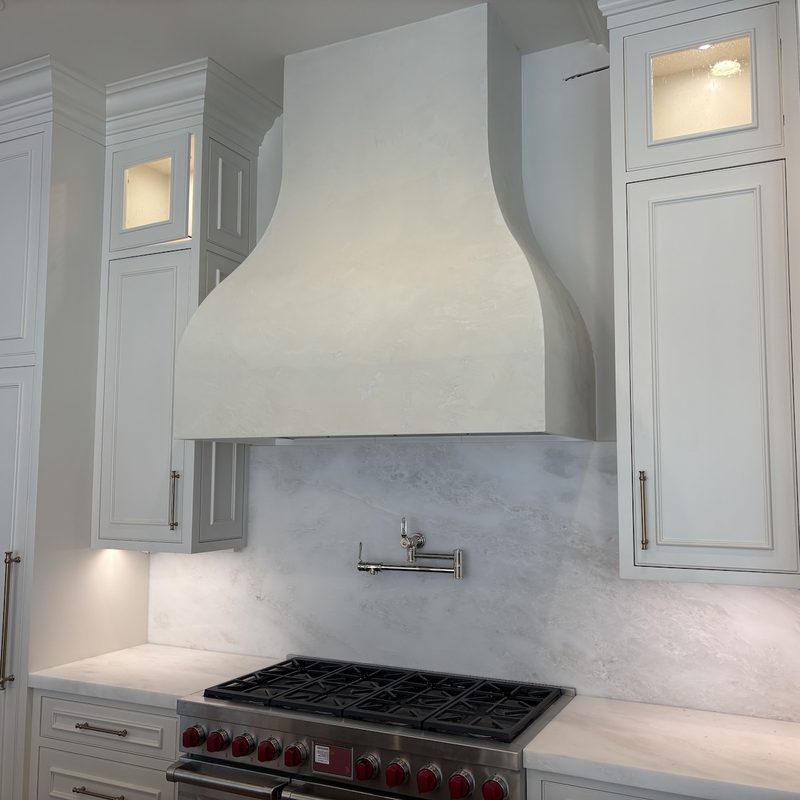 Curved white range hood with a plaster-look finish between glass-front cabinets, above a marble backsplash, pot filler and range