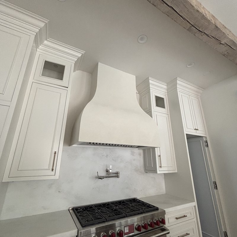 White kitchen with a curved range hood in a soft white plaster look between tall white cabinets, above a marble backsplash and a professional range