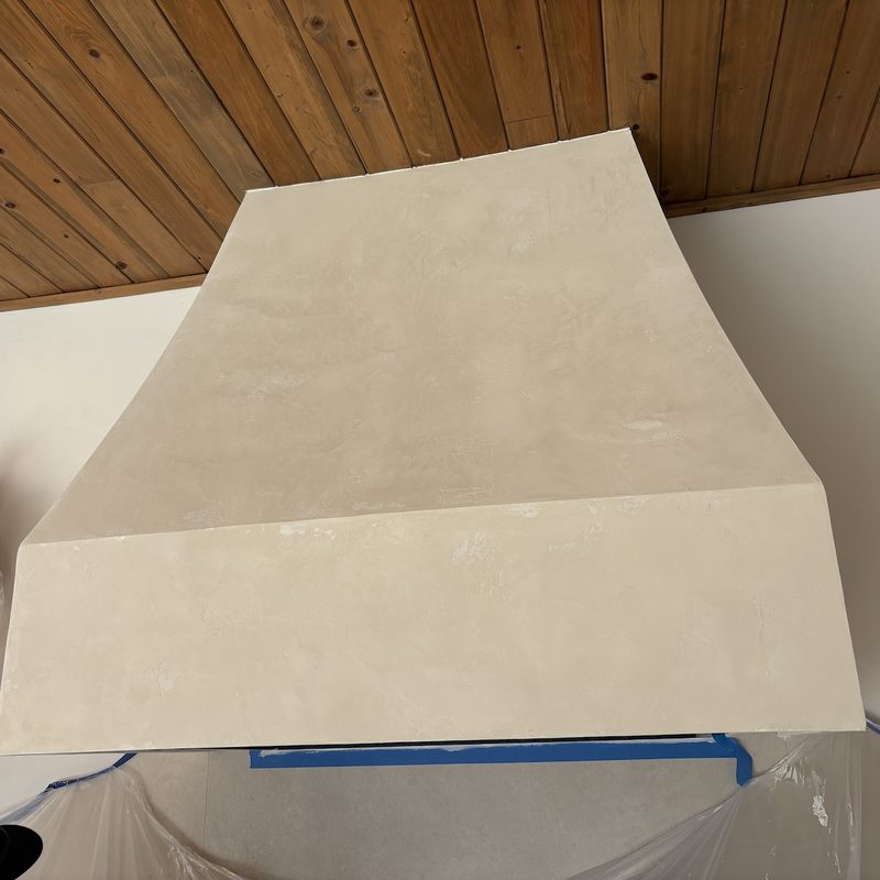Front view of a tapered range hood with a cream, plaster-look finish under a wood plank ceiling, with plastic sheeting below