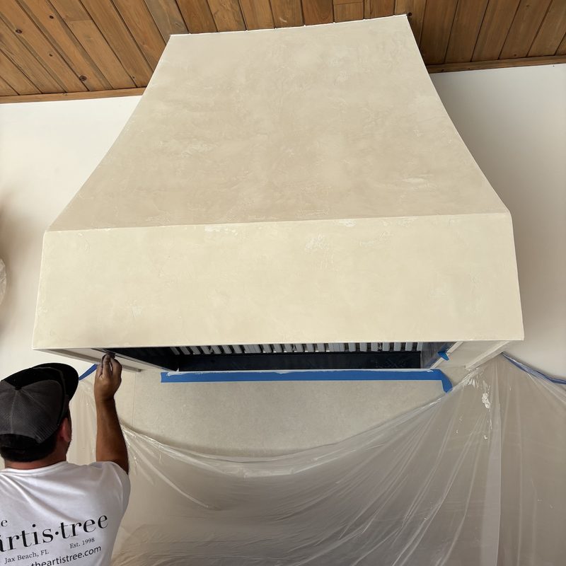 A painter working beneath the tapered cream range hood, with the wall below covered in plastic
