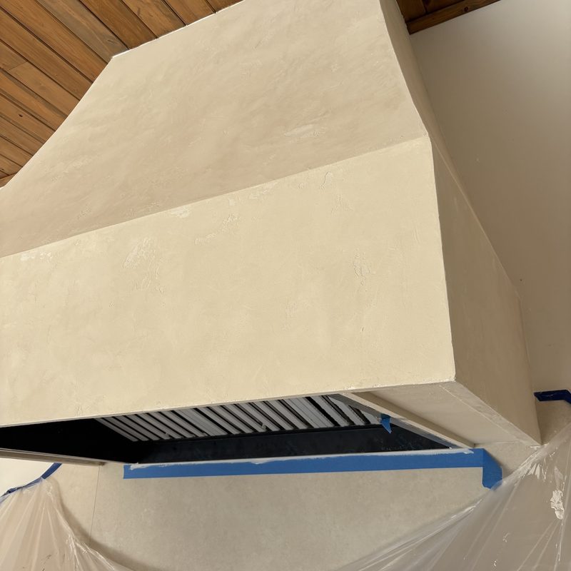 Looking up at the tapered cream range hood under a wood plank ceiling, its lower edge lined with blue painter's tape