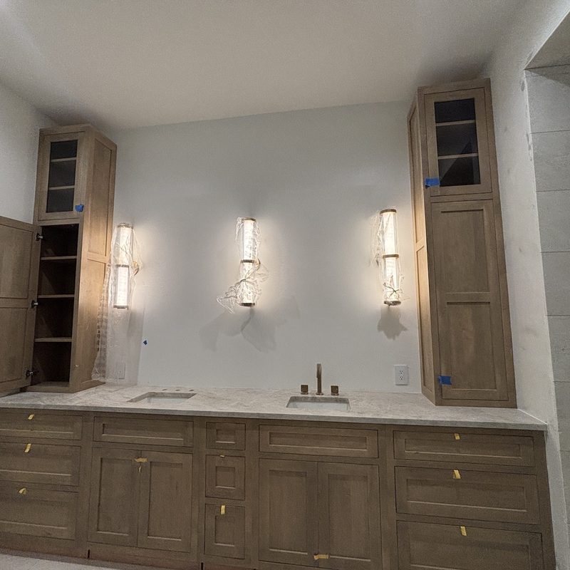 Bathroom vanity wall with three tall sconces between light oak cabinets, walls in a soft white finish