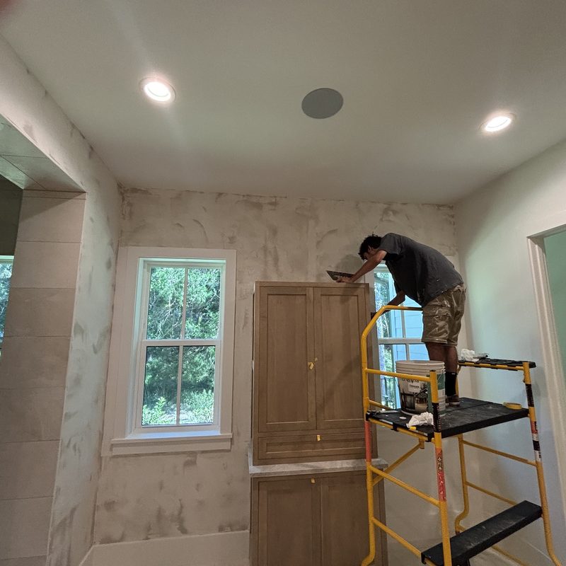 A painter on a rolling scaffold working a mottled white wall finish above an oak cabinet, beside a window