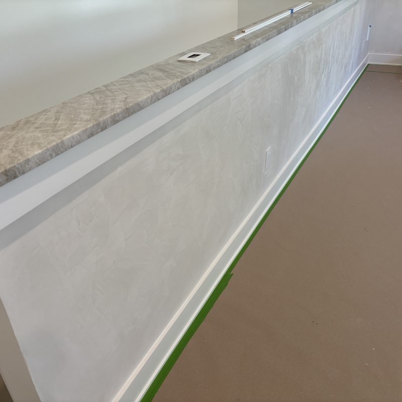 Wainscot-height wall in a soft white, mottled finish under a gray marble-look ledge, with green tape along the baseboard